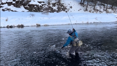 An angler holds their rod high while netting a fish -- river banks covered in snow
