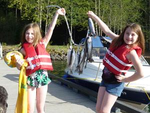 Two girls stand on a dock next to a boat. Each girl is holding up an end of a string of fish they presumably caught