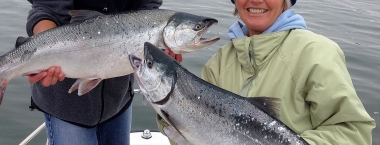 Two people standing on a boat each hold a large silver coho salmon
