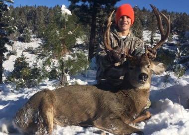 In a snowy field, a rifle hunter shows his large mule deer harvest.