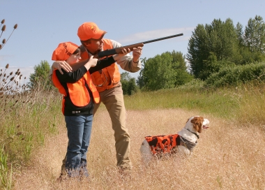 A shooting instructor helps a young student steady her shotgun, as they stand in an open field.