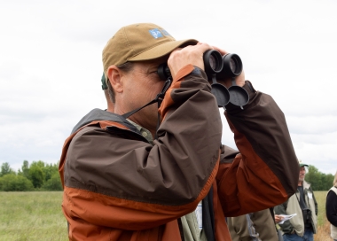 A birder in an open field holds binoculars to his face to look for birds.