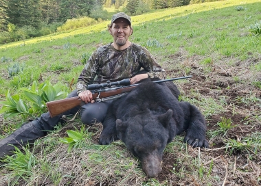 A smiling hunter sits on the ground next to his recently harvested bear.