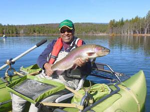 A man in a lime green inflatable pontoon boat holds a large rainbow trout.