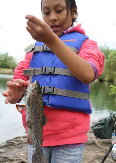 A girl wearing a pink sweatshirt and blue life jacket holds a trout on a line