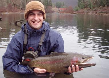 A young man holds a large rainbow trout next to a waterbody