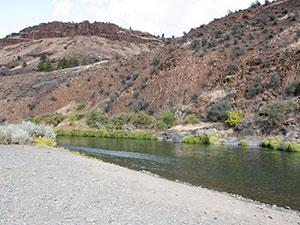 A portion of a creek that runs into the John Day river. The bank is sandy with rocks and on the far side of the river the bank slopes steeply upward 