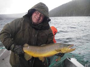 A man stands in a boat holding a large brown trout. The trout is yellowish on the belly and has black speckles on its sides