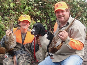 A man and boy hold pheasants they have harvested. Their dog sits between them