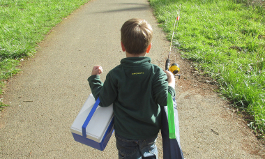 Young boy walking on trail withe arms full carrying cooler, camp chair and fishing rod.
