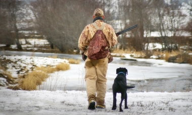A hunter dressed in camo and a black dog walk down a snowy road