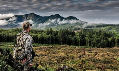 A young man carrying archery equipment looks over a lush forest with mountains in the background and cloudy skies