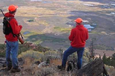 Two hunters in red sweatshirts stand on a high ridge scanning the landscape below