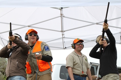 Under a white canopy, two shooters aim shotguns while instructors look on