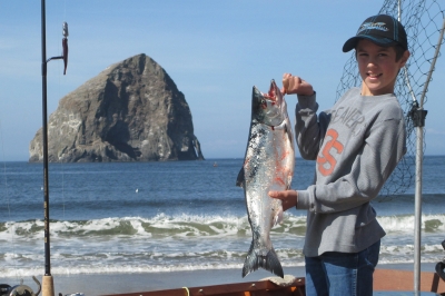 Young angler standing a dory boat holds up coho salmon -- Haystack Rock in background