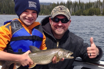 Boy in a boat holds up a lake trout with adult fishing partner giving thumbs up