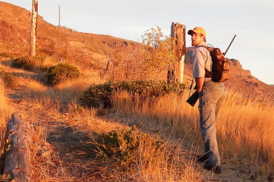 Hunter with rifle on shoulder walks up a dry grassy slope