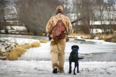 A hunter dressed in camo and a black dog walk down a snowy road