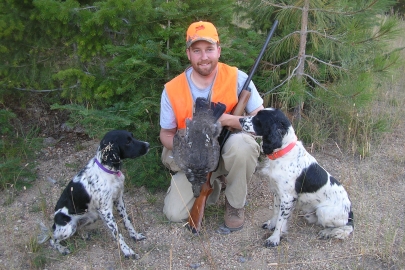 Hunter holding a grouse poses with two black and white hunting dogs