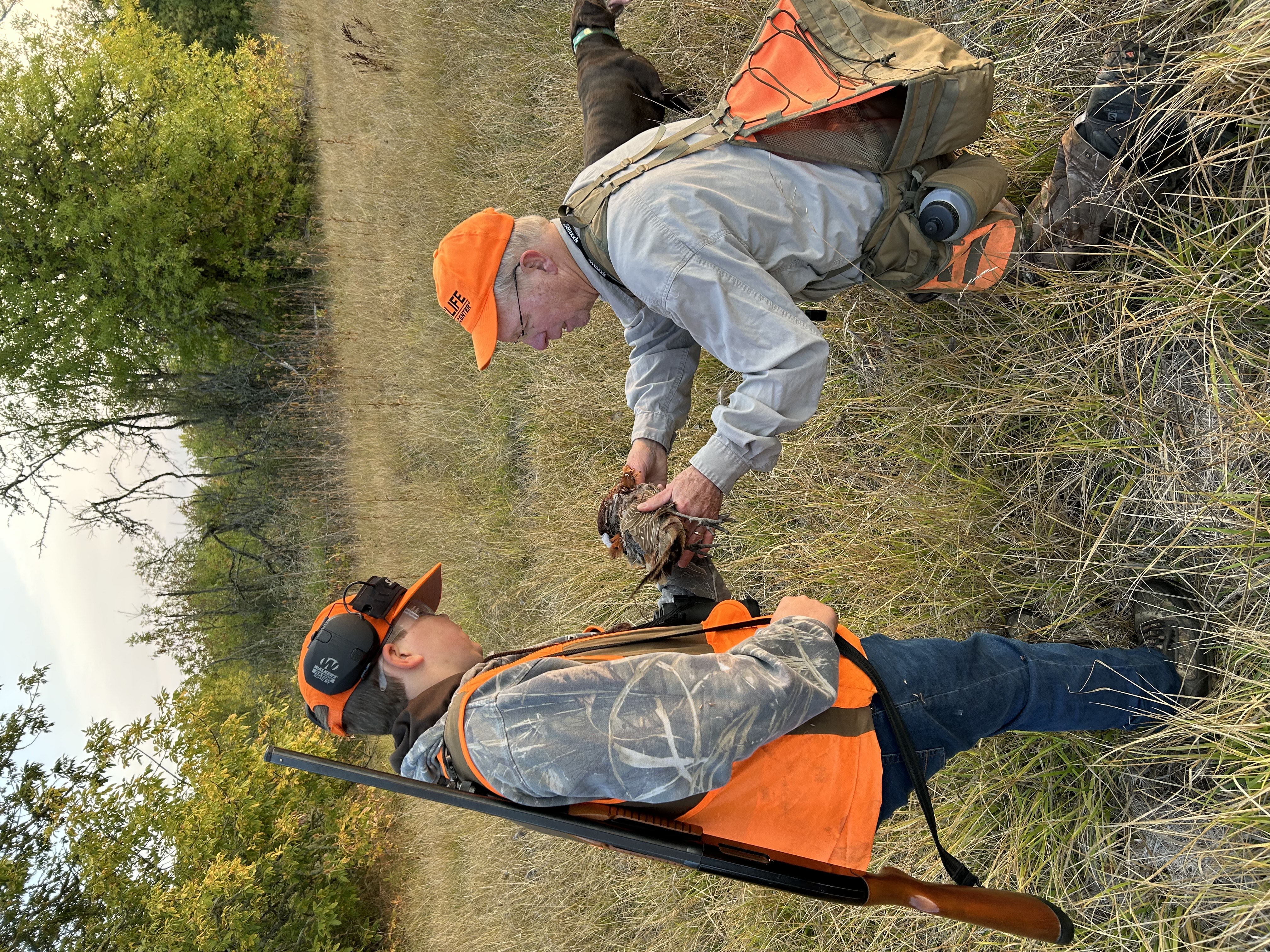 Two individuals wearing bright orange outdoor gear stand in a grassy field with trees in the background. One individual holds a harvested upland game bird while the other looks at it closely. Both carry hunting equipment, including a shotgun and a game vest. A dog is partially visible near the lower edge of the image. The setting appears to be an open, grassy area with scattered shrubs and soft light.